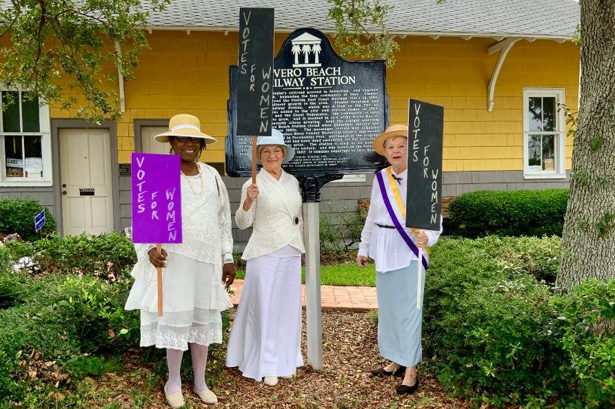 Ladies holding "Vote for Women" signs in front of Vero Beach Railway Station historical sign